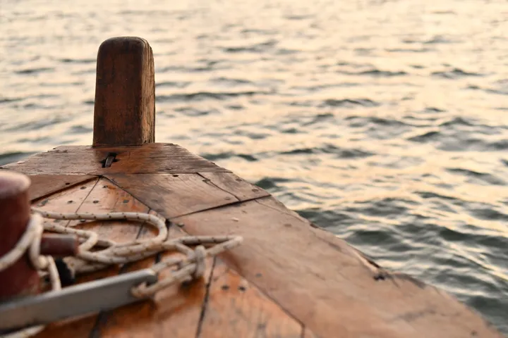 Le pont avant d’une barque en bois, une bitte d’amarrage usée et un cordage blanc lové, devant une eau argentée par la fin du jour.