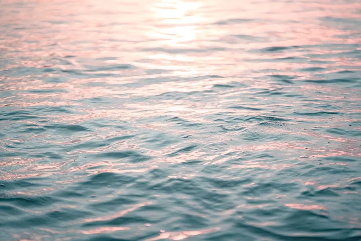 La surface d’une mer à peine ridée, où le couchant pose de larges reflets rose pêche sur une eau vert d’eau.